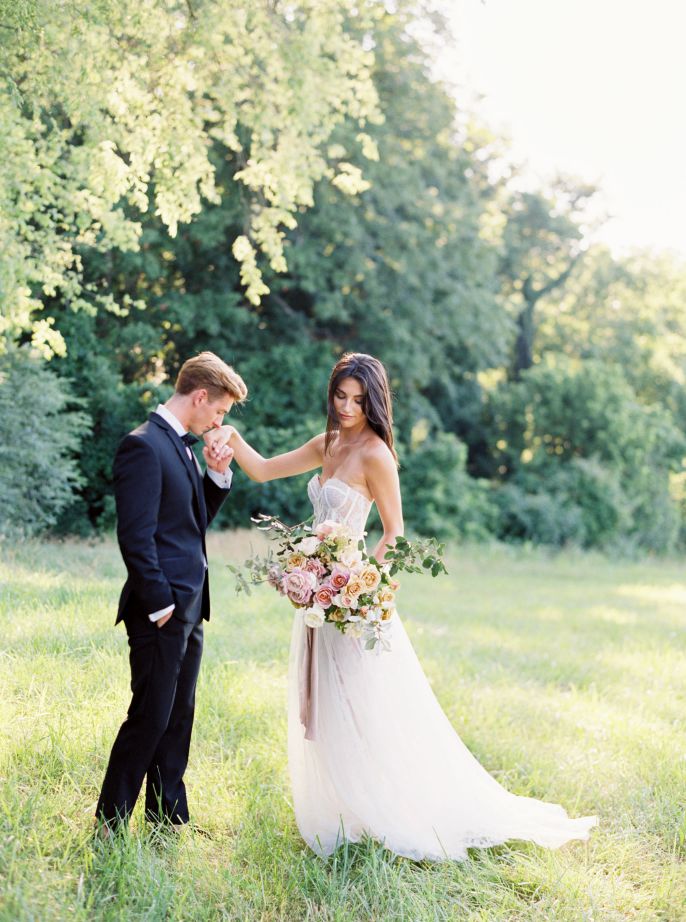 groom kissing bride's hand in grassy field
