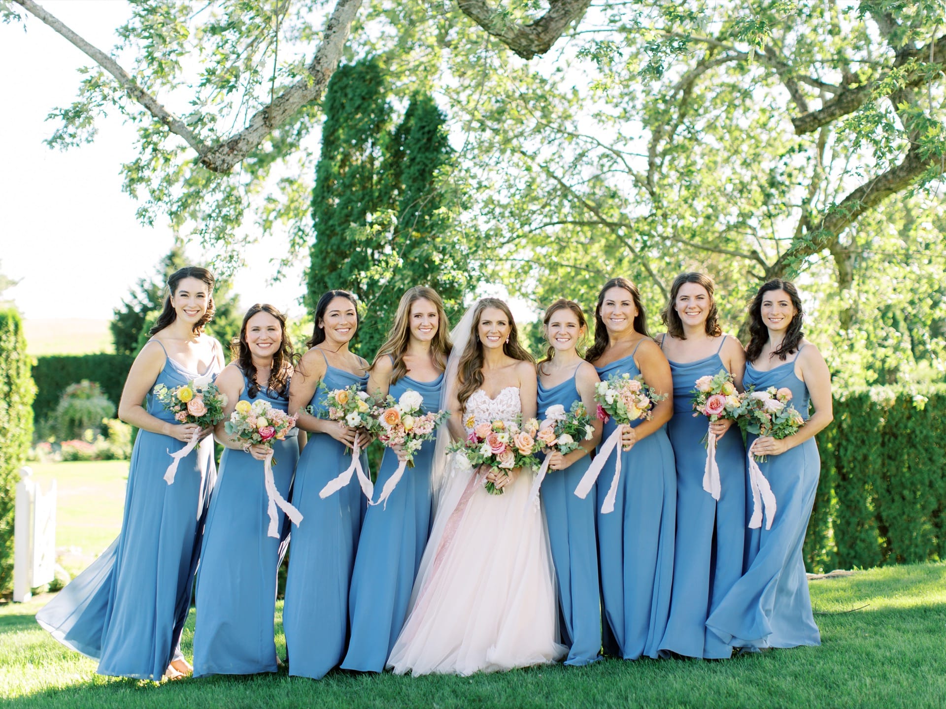 Bride in white surrounded by bridesmaids in blue dresses, standing in front of green hedges and trees