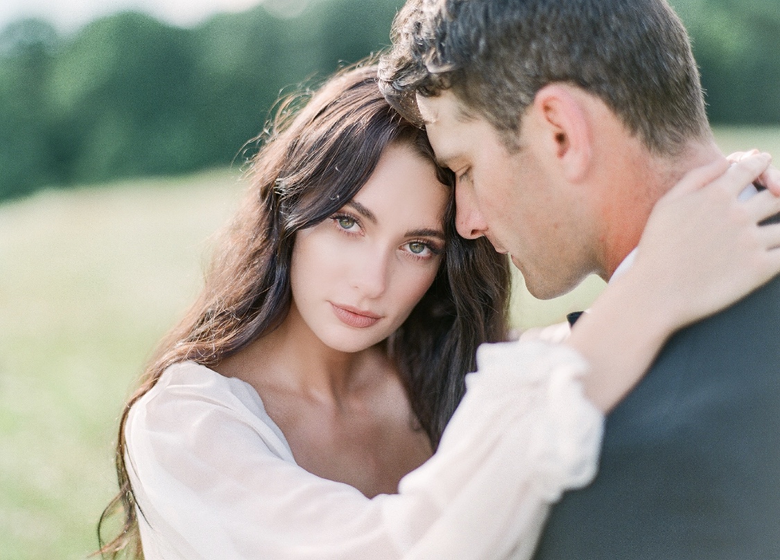 Bride with elegant brown hair looking into the camera as groom looks over her shoulder, standing in a field