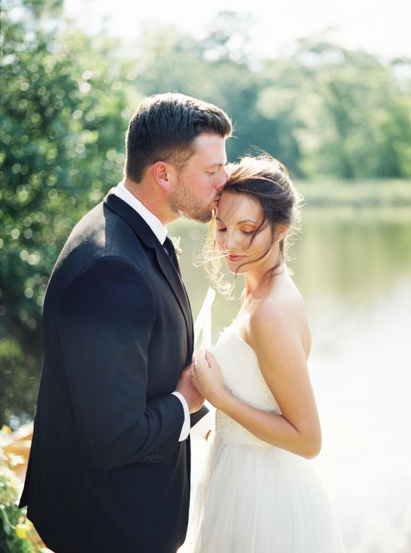 groom kissing bride's forehead in front of a lake