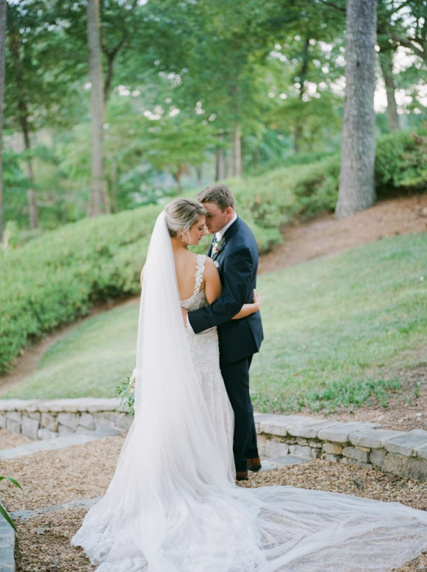 Bride and groom softly embrace on a path in front of green grass.